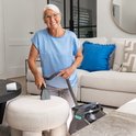 Een oudere vrouw met bril reinigt met een grijze reinigingsmachine een witte poef in een woonkamer.