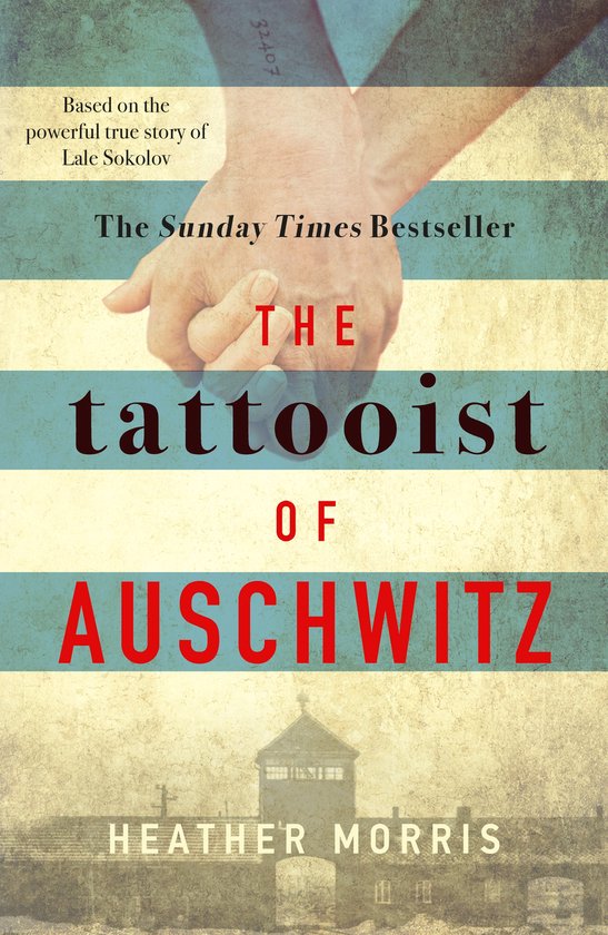 Boekomslag van 'The Tattooist of Auschwitz'. Twee handen houden elkaar vast, met een tatoeagenummer op de arm. De titel is groot en rood en blauw gedrukt.