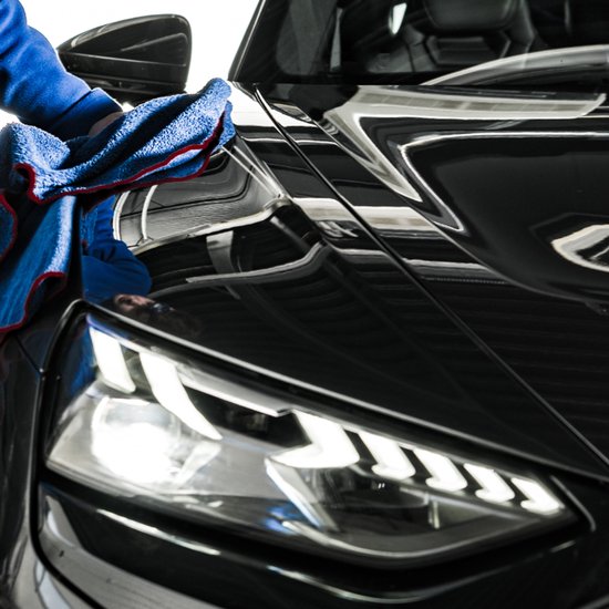 Een hand met een blauwe microvezeldoek poetst de glimmende zwarte motorkap van een auto, de koplampen zijn zichtbaar.