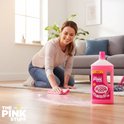 Vrouw met roze doekje maakt de vloer schoon in woonkamer, naast een fles The Pink Stuff vloerreiniger.