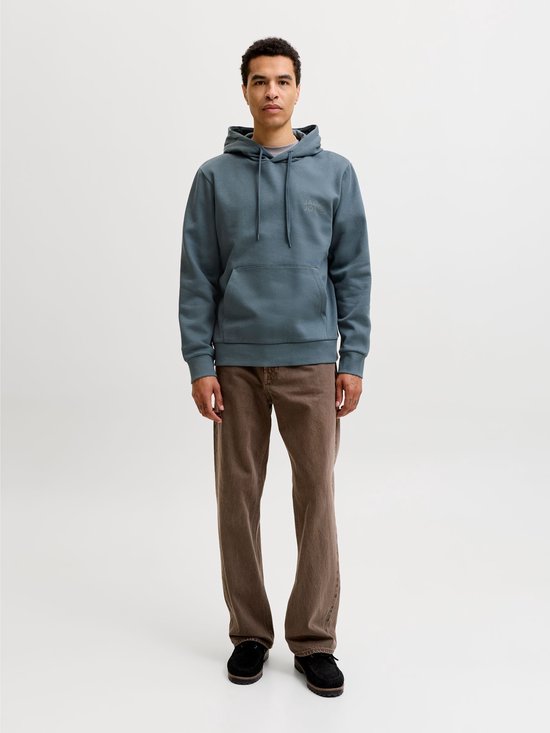 Man staat recht voor een witte achtergrond, gekleed in een blauwgrijze hoodie en bruine broek.
