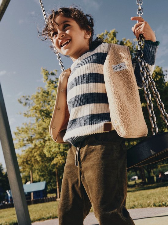 Jongen met krullend haar op een schommel, draagt een beige teddy gilet over een gestreepte trui.