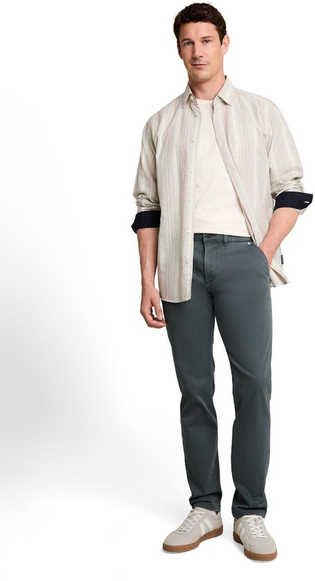 Model draagt een donkergrijze chino, gestreept overhemd, beige T-shirt en lichte sneakers.