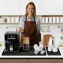 Zwarte koffiebarmat met een zwarte volautomatische espressomachine, een zak koffiebonen, een witte waterkoker, twee witte melkkannetjes en twee espressokopjes.