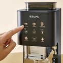 Vinger drukt op "ESPRESSO" op het bedieningspaneel van de zwarte Krups koffiemachine.