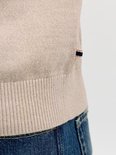 Onderlichaam van een man in een lichtbeige gebreid poloshirt met lange mouwen en geribde zoom, met een donkerblauwe jeans en een stoffen label met rode en blauwe strepen.