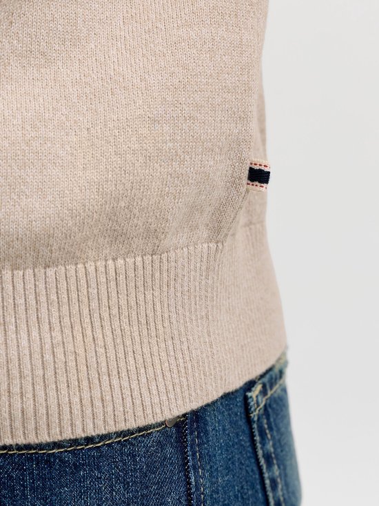 Onderlichaam van een man in een lichtbeige gebreid poloshirt met lange mouwen en geribde zoom, met een donkerblauwe jeans en een stoffen label met rode en blauwe strepen.