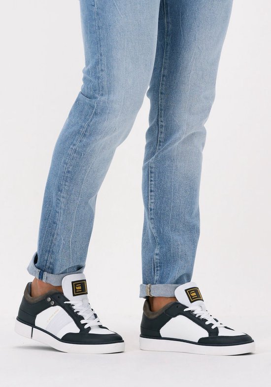 Model draagt witte sneakers met marineblauwe en bruine accenten, gecombineerd met een spijkerbroek.