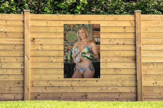 Blonde vrouw in kleurrijke bikini schenkt drank bij een bar, opgehangen aan houten schutting.