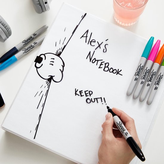 Een hand schrijft "KEEP OUT!!" op een notitieboek met een zwarte Sharpie stift. Stiften liggen ernaast.