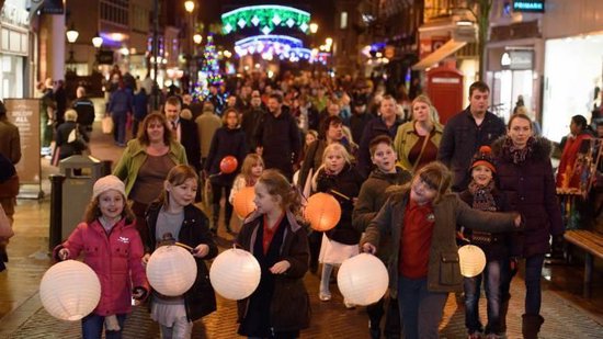 Kinderen lopen met witte papieren lampionnen door een winkelstraat in de avond.