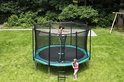 Een zwart trampoline net met een groene rand, met twee kinderen erop en ernaast, in een tuin.