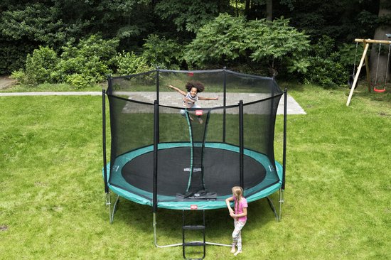 Een zwart trampoline net met een groene rand, met twee kinderen erop en ernaast, in een tuin.
