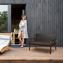 Vrouw in zomerkleding staat naast een donkerbruine tuinbank op een houten terras.