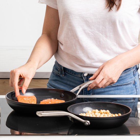 Persoon legt stukjes zalm in een zwarte KitchenAid-pan naast een pan met geroosterde hazelnoten.