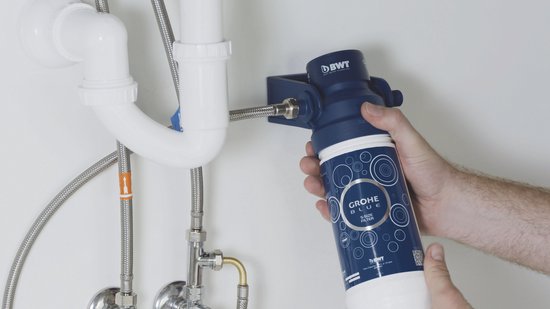 Handen bevestigen een blauwe GROHE BLUE S-SIZE waterfilter aan een gootsteenafvoerbuis.