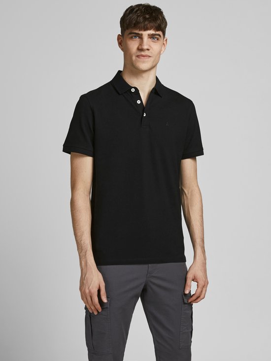 Man draagt een zwarte polo met knopen en een geborduurd logo, en een grijze broek.
