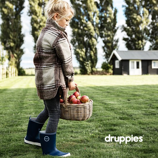 Meisje met blonde paardenstaart loopt door een grasveld, draagt een geruite jas, grijze jeans en donkerblauwe rubberlaarzen, met een mand met appels.