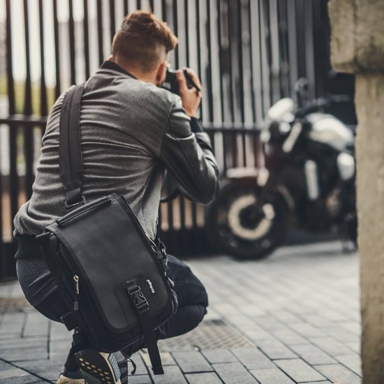 Zwarte Kriega sling tas gedragen door een man die knielt om een motorfiets te fotograferen.
