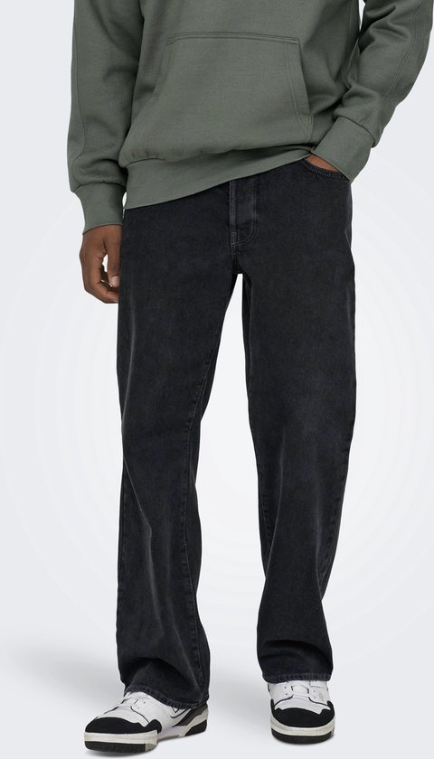 Man met de handen in de zakken van een wijde, zwarte spijkerbroek met stonewashed effect, met groene hoodie en sneakers.