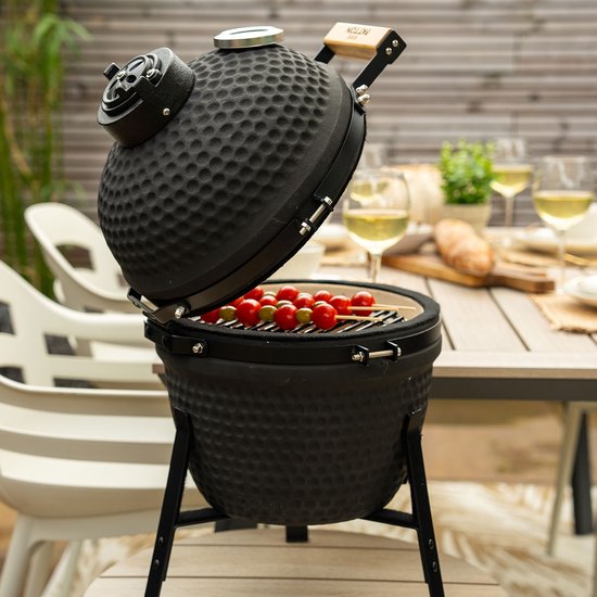 Zwarte kamado barbecue met open deksel op een tuintafel, met spiesjes met tomaten en mozzarella.