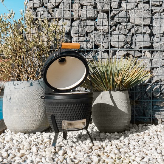 Zwarte kamado barbecue met open deksel, grillrooster en houten handvat, in een tuin.