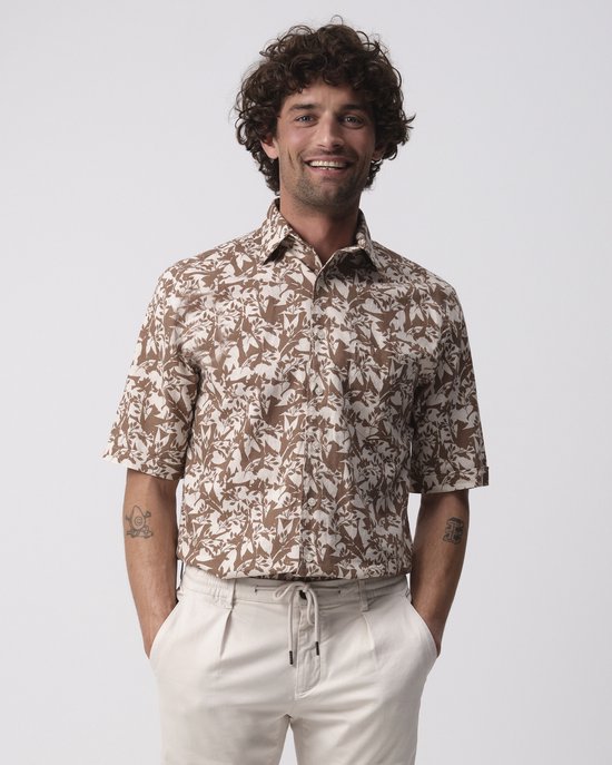 Man met krullend bruin haar, draagt een bruin overhemd met bladerpatroon en een beige broek.