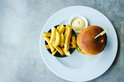 Een goudbruine burger op een wit bord, naast een kom met friet en een bakje mayonaise.