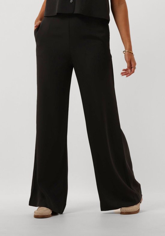 Voor- en zijkant van een zwarte wijde broek, gedragen door een model met sieraden.