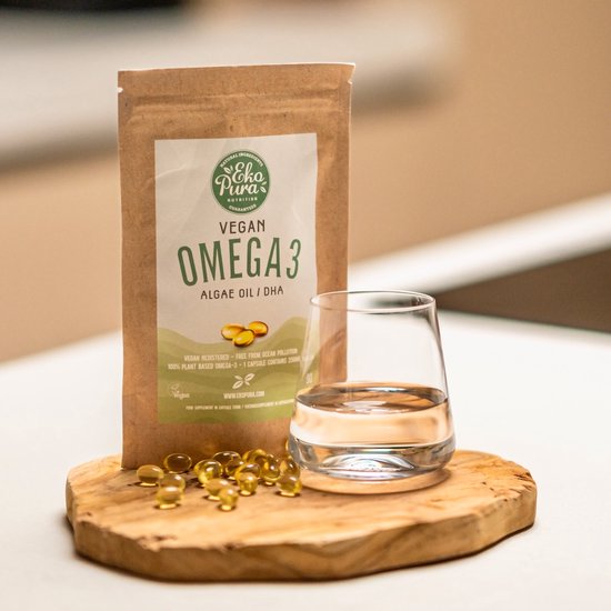 Bruine papieren zak van 'Ekopura Vegan Omega 3 Algae Oil' en goudkleurige capsules naast een glas water op een houten plank.