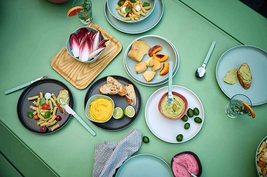 Een bovenaanzicht van een tafel vol eten, met salades, brood, dips en fruit. Diverse lichtgroene en grijze schalen.