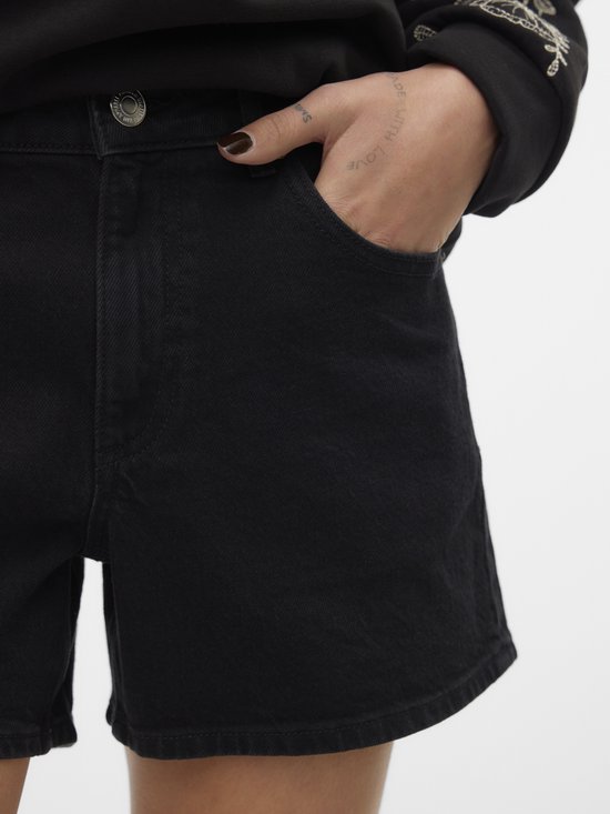 Model met hand in zak van zwarte denim shorts. Zwarte trui met geborduurde mouwen.