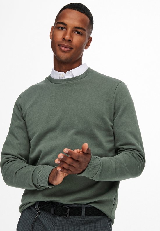 Man kijkt naar de camera, handen tegen elkaar. Draagt een groene sweater, wit overhemd eronder en grijze broek met riem.