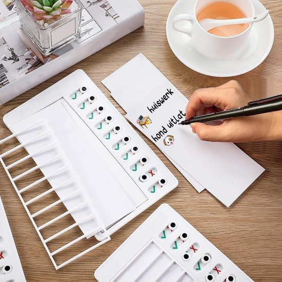 Witte planborden op een houten tafel. Een hand schrijft taken op een notitievel. Met schuifjes voor vinkjes en kruisjes.