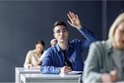Student met bril steekt hand op in een klaslokaal, schrijft met pen.