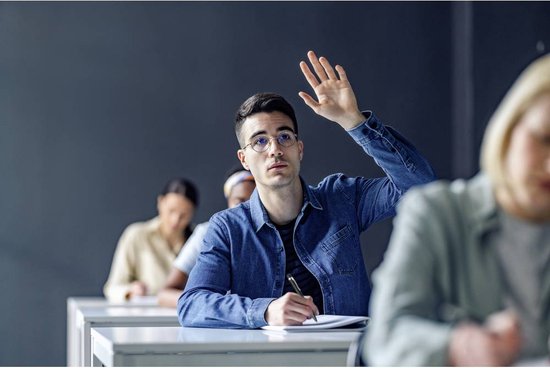 Student met bril steekt hand op in een klaslokaal, schrijft met pen.