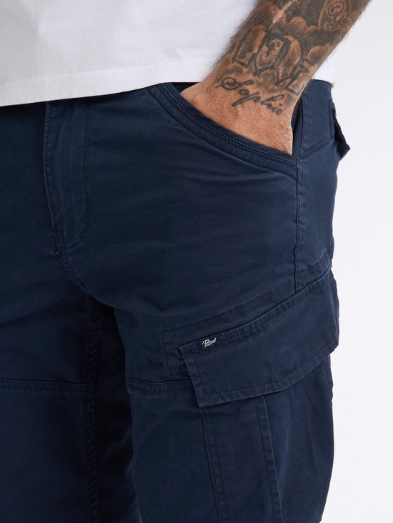 Donkerblauwe cargobroek met klepzak op het been, zichtbare tatoeage op de arm.