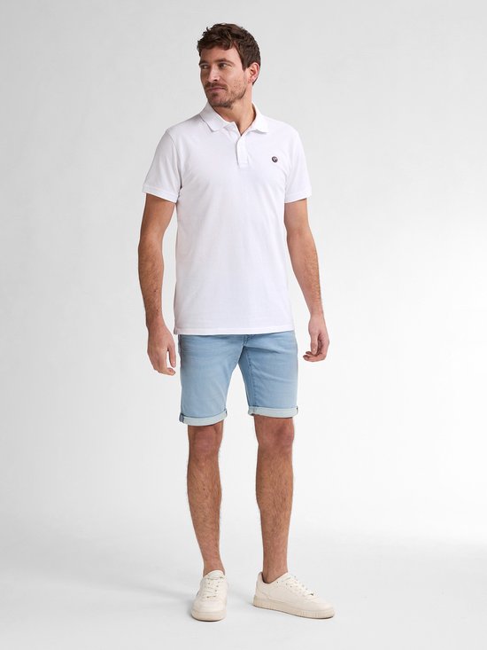 Een witte polo met een klein logo op de linkerborst, gedragen door een man met een lichte spijkerbroek.