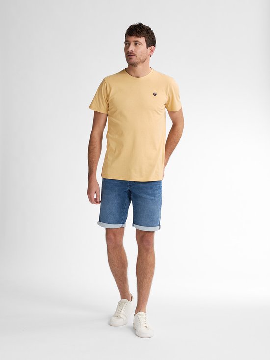 Man met licht getinte huid draagt een donkergeel T-shirt, blauwe spijkerbroek, witte sneakers en witte sokken.