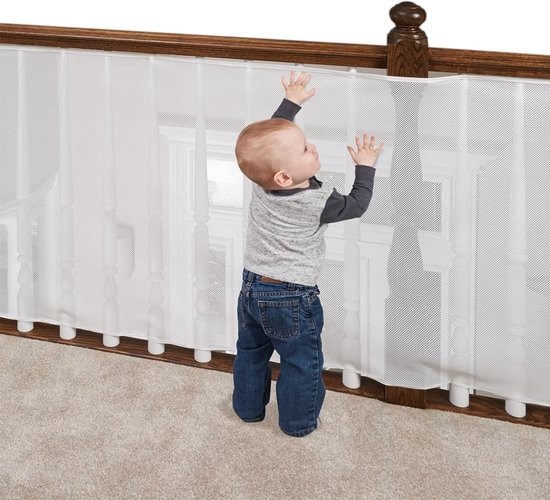 Een baby in spijkerbroek en shirt reikt naar een wit gaashekwerk langs een houten trapleuning.
