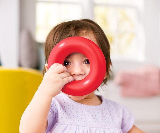 Een baby met bruin haar houdt een rode ring van de Fisher-Price stapelpiramide voor haar gezicht. Ze glimlacht.