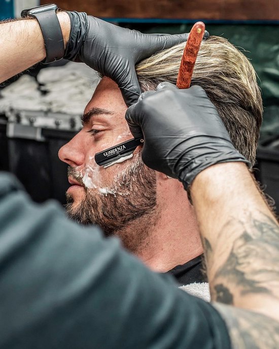Barbier met zwarte handschoenen scheert een man met het scheermes met houten handvat. De man heeft blond haar en een baard.