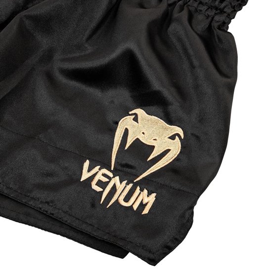 Detailopname van het goudkleurige Venum-slangenlogo en tekst op de zwarte satijnen broekspijp.
