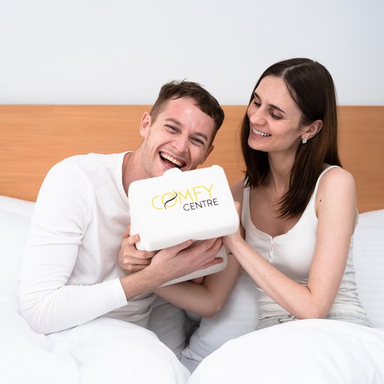 Lachend stel op een bed, vasthoudend een wit Comfy Centre kniekussen.