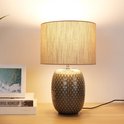 Verlichte grijze tafellamp met beige kap en decoratieve voet op een houten tafel.