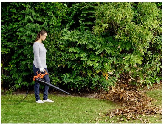Vrouw in grijze trui en spijkerbroek gebruikt oranje bladblazer op gras naast heg.