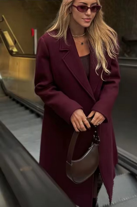 Vrouw op roltrap in een bordeauxrode jas met reverskraag, zonnebril en bruine tas.