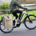 Beige dubbele fietstas aan de achterkant van een elektrische fiets, bereden door een persoon in een groene jas.