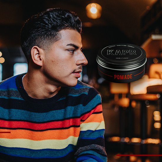 Jonge man met gestyled haar, draagt een veelkleurige trui. Naast hem staat een blikje KAPR Pomade.