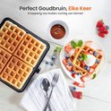 Vier goudbruine wafels in een open wafelmaker, met een bord met daarop wafels, slagroom, fruit en blauwe bessen. Een kom met chocoladesaus staat ernaast.